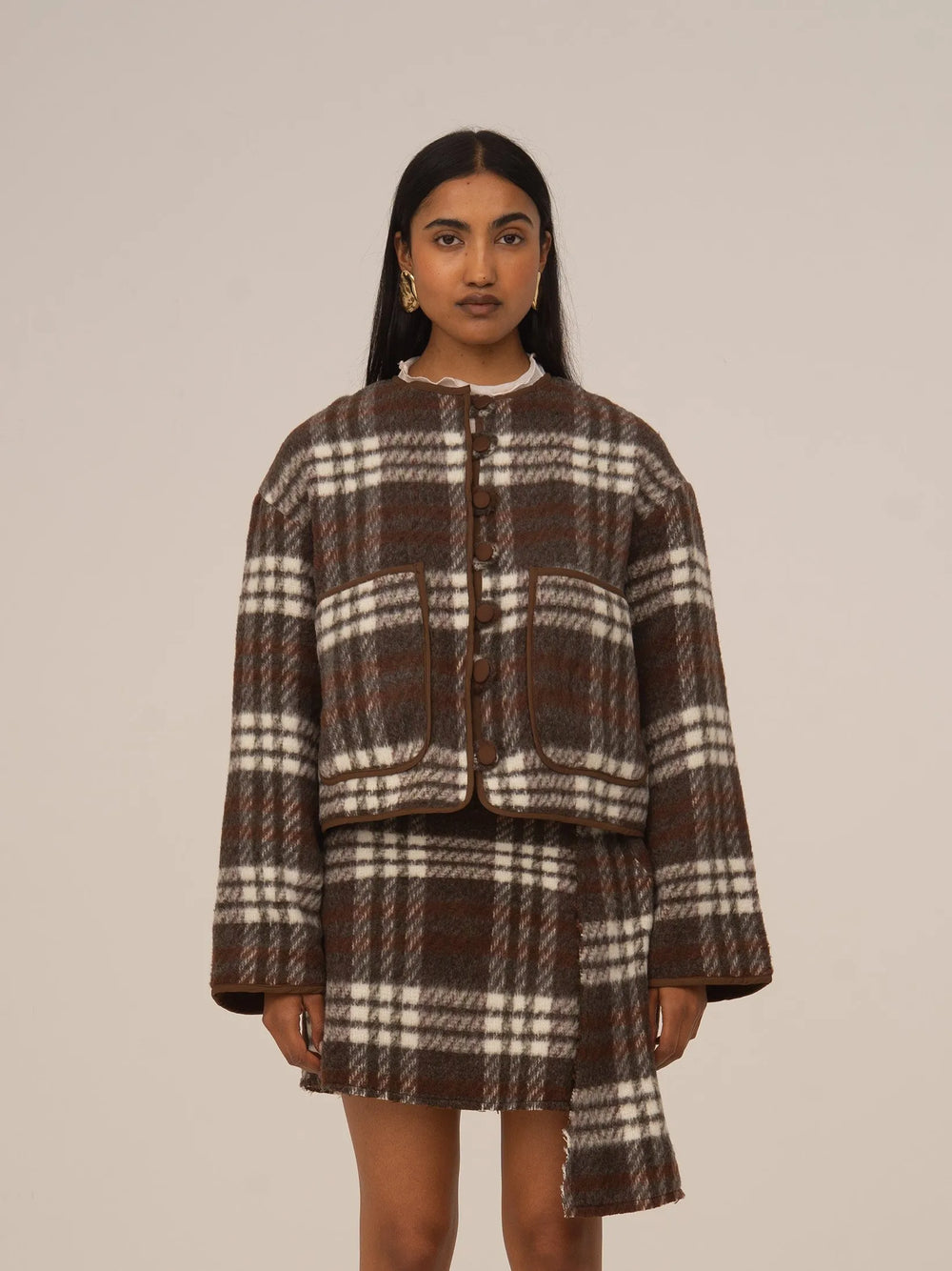 Chaqueta corta de cuadros marrón con botones frontales y cuello redondo, combinada con falda a juego, presentada sobre modelo en estudio con fondo neutro.