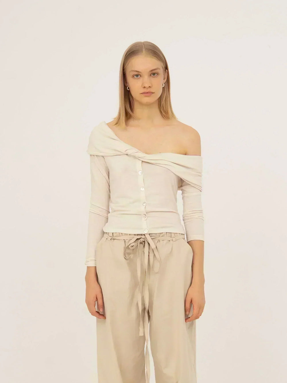 Top blanco de punto con diseño off-shoulder, detalle drapeado cruzado en el escote y cierre frontal con botones. Silueta ajustada y manga larga, confeccionado en jersey suave para un ajuste cómodo y femenino.