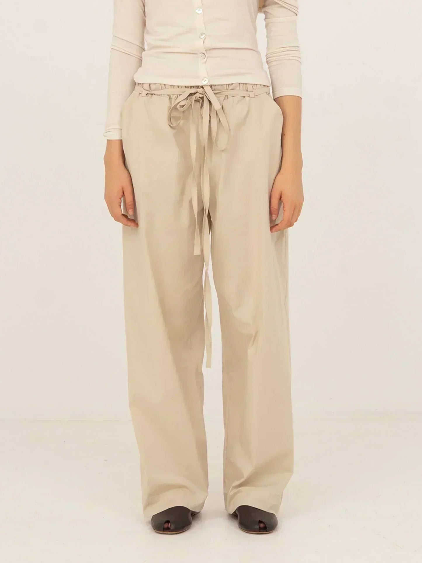 Pantalón beige de tejido fluido con corte recto y pernera larga, cintura elástica con lazada ajustable y caída suave, presentado sobre modelo en estudio con fondo neutro.