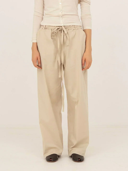 Pantalón beige de tejido fluido con corte recto y pernera larga, cintura elástica con lazada ajustable y caída suave, presentado sobre modelo en estudio con fondo neutro.