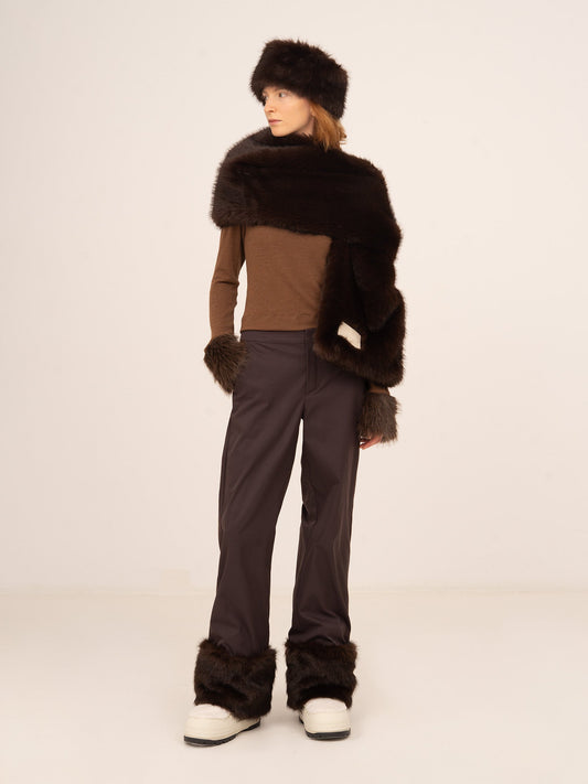 Modelo femenina vistiendo pantalón marrón oscuro con detalles de pelo en el bajo, combinado con top en tonos tierra y accesorios de pelo, fotografiada de cuerpo entero sobre fondo neutro.
