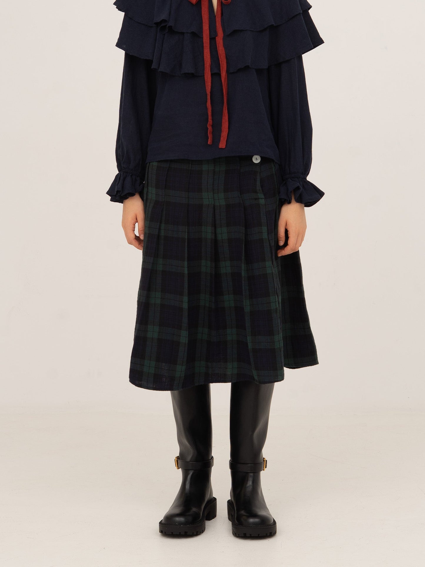 Falda midi plisada de cuadros en tonos azul y verde con detalles en rojo, silueta recta y caída fluida, combinada con botas negras y presentada sobre modelo en estudio con fondo neutro.