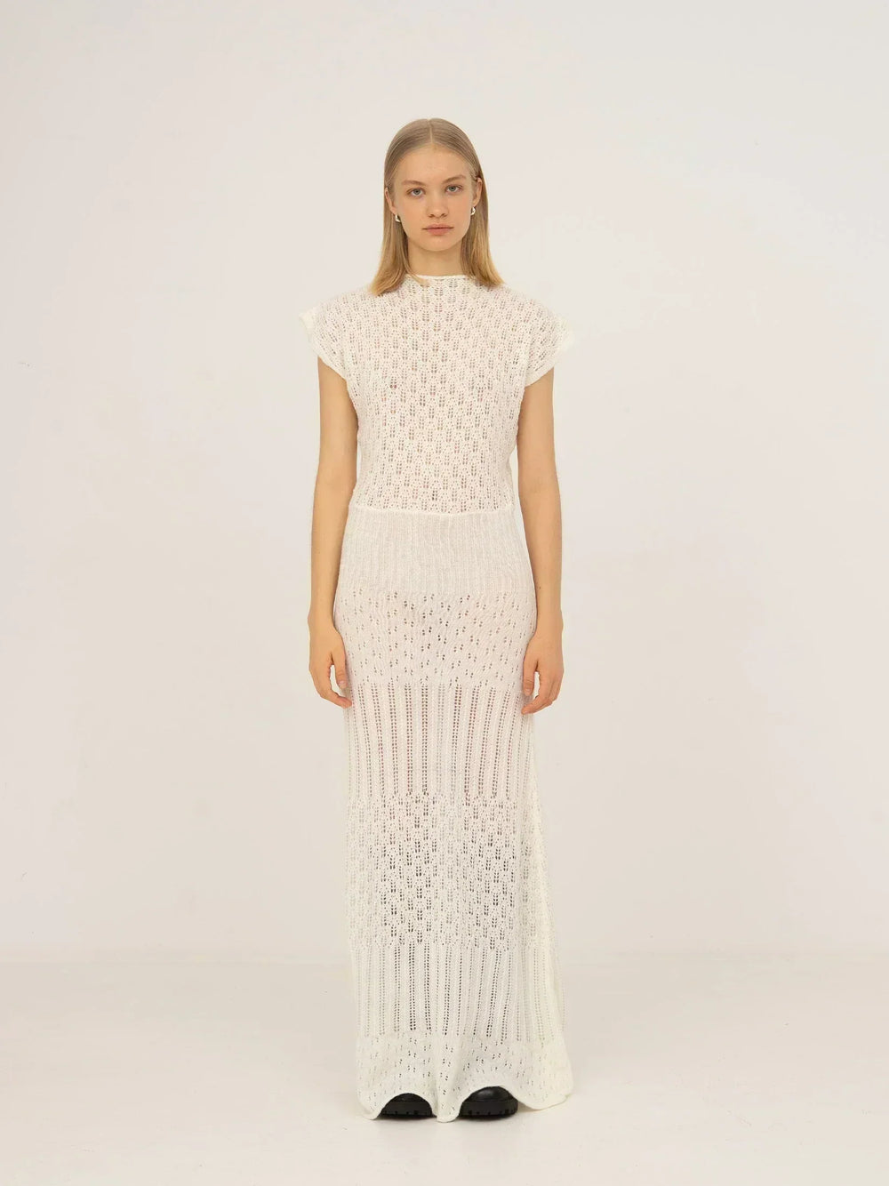Vestido maxi blanco de punto calado con diseño de textura abierta, cuello alto, manga corta y silueta recta de caída ligera, presentado sobre modelo en estudio con fondo neutro.