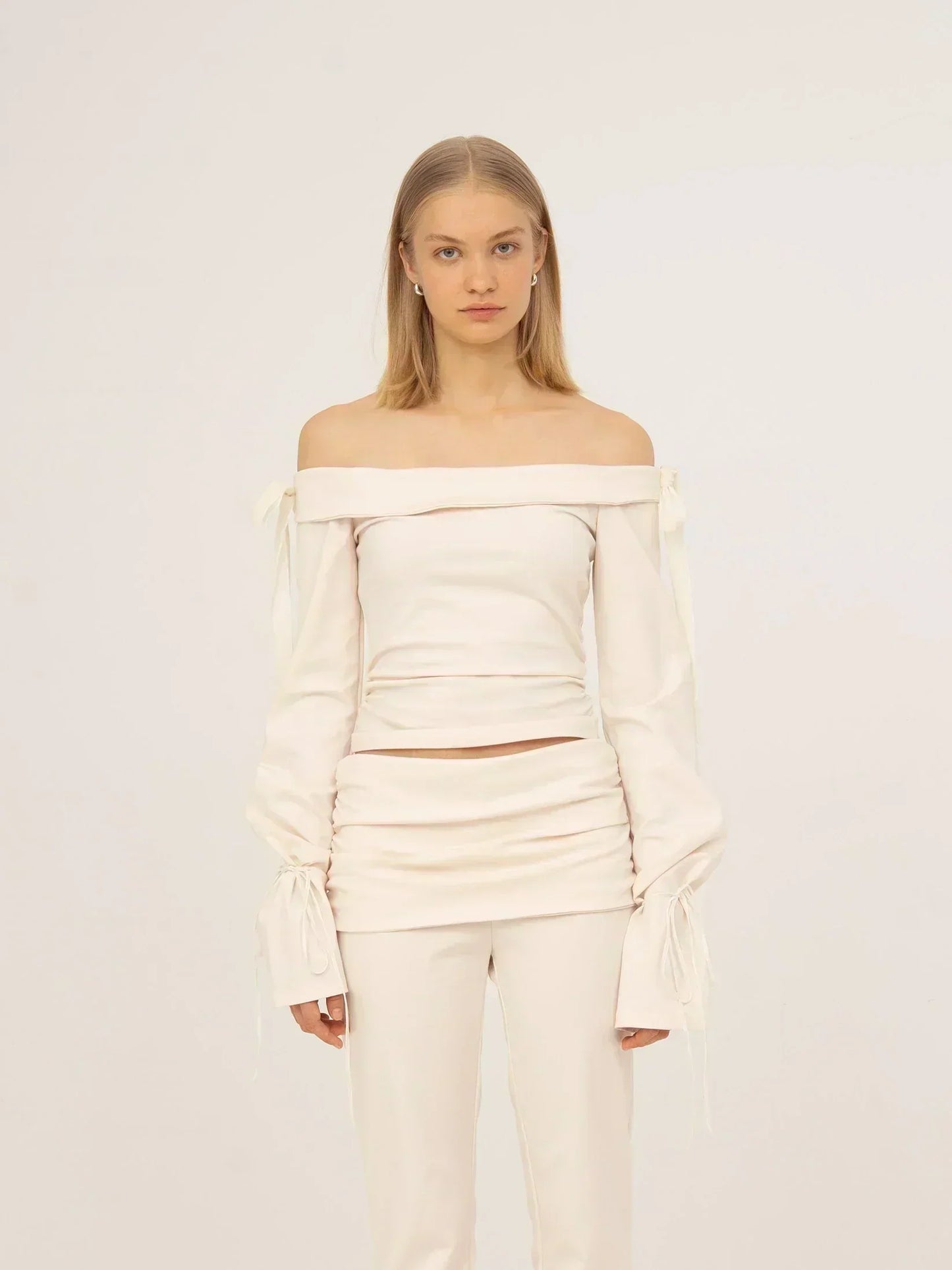 Top blanco off-shoulder de tejido jersey con escote bardot, mangas largas con detalle fruncido y lazadas en los puños, silueta ajustada y acabado limpio, presentado sobre modelo en estudio con fondo neutro.
