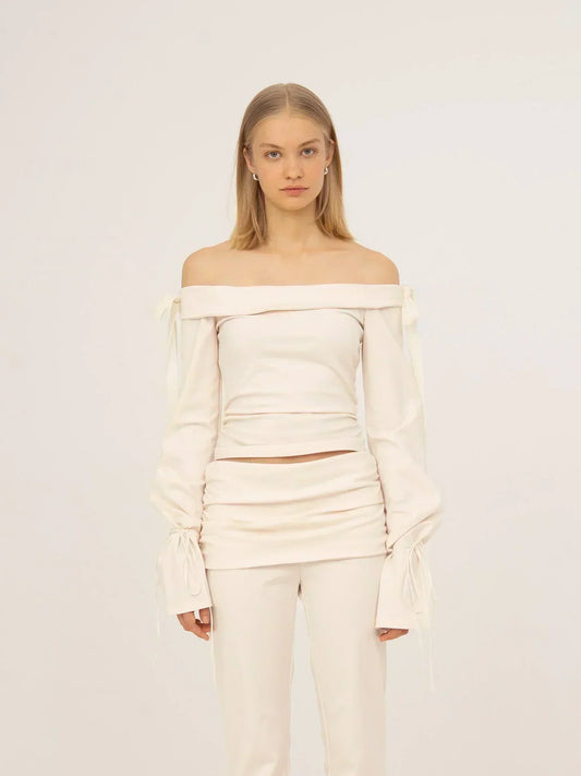 Top blanco off-shoulder de tejido jersey con escote bardot, mangas largas con detalle fruncido y lazadas en los puños, silueta ajustada y acabado limpio, presentado sobre modelo en estudio con fondo neutro.